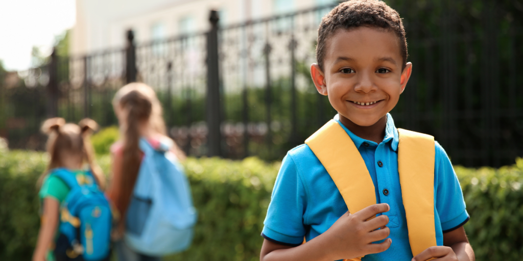 Young boy wearing a blue shirt, yellow backpack, and a big grin in front of a school building.